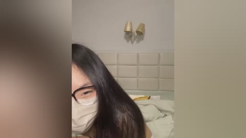 Media: Video of a woman with long black hair and glasses, wearing a mask, partially visible in a dimly lit bedroom with beige walls and a white padded headboard.