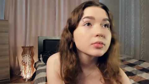 Media: A video of a young woman with long brown hair, wearing a strapless top, sitting on a bed with a striped blanket. She has a neutral expression.