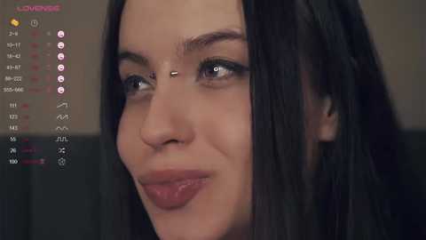 Media: Close-up video of a young woman with straight, dark brown hair, light skin, and a subtle smile, looking to the right. Background includes a blurred, dark sofa and a digital display showing time, date, and weather information.