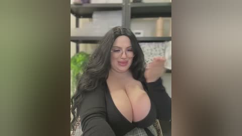 Media: A video of a voluptuous, fair-skinned woman with long, wavy black hair and glasses, wearing a plunging black dress, standing in a modern office with bookshelves and greenery in the background.