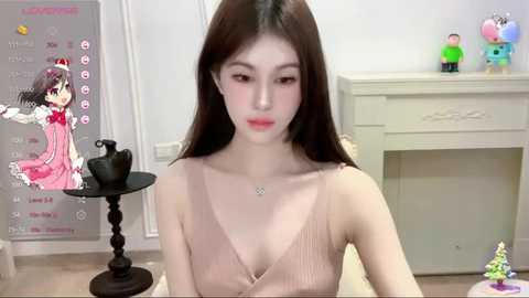 Media: Video of a fair-skinned Asian woman with long dark hair, wearing a beige sleeveless dress, seated indoors. Background includes a calendar, figurines, and a black table.