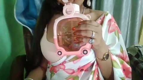 Media: Video of a woman with long black hair wearing a white blouse and floral-patterned dress, holding a pink baby bottle with a star-shaped nipple.