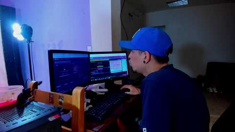 Media: A video of a young man with a light complexion, wearing a blue cap and headphones, sitting in a dimly-lit room, focused on a computer screen.