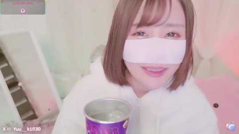 Media: A video of a young Asian woman with shoulder-length brown hair, wearing a white mask and robe, holding a purple cup with ice. She smiles slightly, in a dimly lit, soft-colored room.