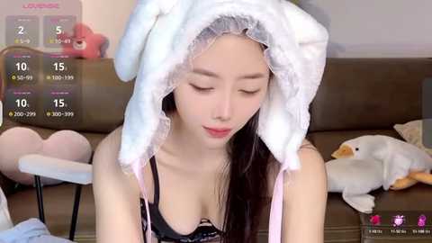 Media: Video of an East Asian woman in a fluffy white bunny headband, wearing a black bra, sitting on a brown couch. Background features plush toys, a cat cushion, and a virtual game overlay.