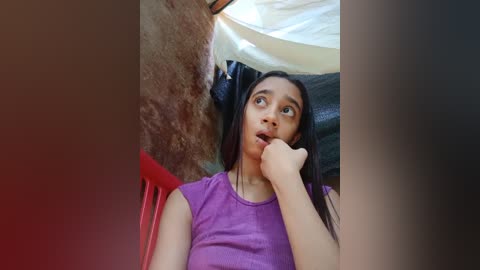 Media: Video of a young girl with light brown skin, long black hair, and wide eyes, sitting on a red chair, looking curious inside a tent with beige and brown fabric.