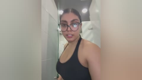 Media: Video of a young woman with dark hair, glasses, and a black tank top, standing in a narrow, dimly lit corridor.