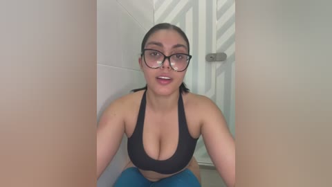 Media: A video of a Latina woman with light brown skin, wearing glasses and a black sports bra, sitting in a bathroom with white tiled walls and a beige door.