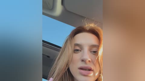 Media: Video of a young woman with fair skin and long, straight, light brown hair, wearing minimal makeup. She has a neutral expression, slightly tilted head, and is captured from a low angle inside a car.