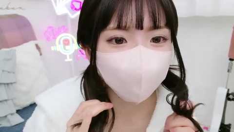 Media: Video of an Asian woman with long black hair, wearing a white face mask, holding her hair in a cozy, possibly bedroom setting with soft pink neon signs.