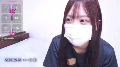 Media: A video of a young East Asian woman with long, straight, dark hair, wearing a black top and a white surgical mask. She appears to be indoors, with a digital thermometer displaying her temperature.