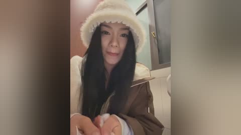 Media: Video of an East Asian woman with long black hair, wearing a white fur hat and a brown jacket, standing in a dimly lit hallway with white walls and a door.