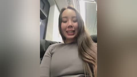 Media: Video of a young Asian woman with long brown hair, wearing a light gray top, smiling while seated in a car. Background shows part of the car interior and a window.