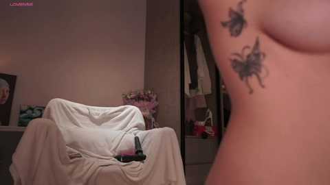 Media: A video shows a nude woman's side profile with butterfly tattoos on her rib cage. She sits on a white chair in a dimly lit bedroom, with a gray wall, a mirror, and a bouquet of flowers in the background.