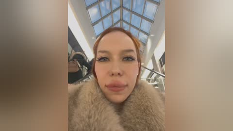 Media: Video of a young woman with fair skin and red hair, wearing a fur coat, standing in a modern indoor setting with glass ceilings and metallic railings.