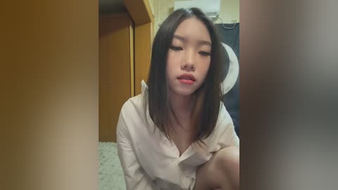 Media: Video of a young Asian woman with straight black hair, fair skin, and full lips, wearing a light-colored shirt. She sits on a bed in a room with wooden furniture, a ceiling fan, and a carpeted floor.