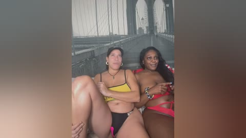 Media: Video of two women sitting on a bridge; one in a yellow bikini, the other in red lingerie, with a blurred cityscape background.