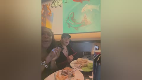 Media: A video of three women in a casual restaurant, one holding a camera. They are seated at a table with a plate of food, surrounded by colorful wall art and a large green screen.