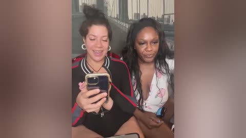 Media: Video: Two women seated together, one with curly hair and a black jacket, the other with long straight hair and floral top, smiling at a phone.