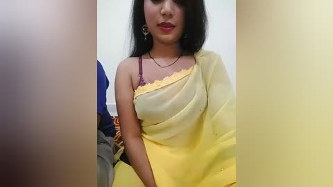 Media: Video of a South Asian woman with medium-dark skin, straight black hair, and red lipstick, wearing a yellow saree and a maroon bra, seated indoors against a plain white background.