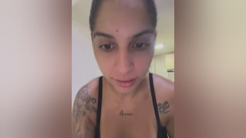 Media: Video of a young, light-skinned woman with dark hair, wearing a black tank top, showing off tattoos on her arms. She has a small mole on her forehead. The background is a blurred indoor setting with neutral tones.