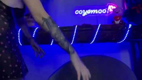 Media: Video of a woman with dark hair, wearing a polka-dotted dress, holding a stuffed animal. Neon sign in background reads \"oyeyemool\" with a glowing blue hue.