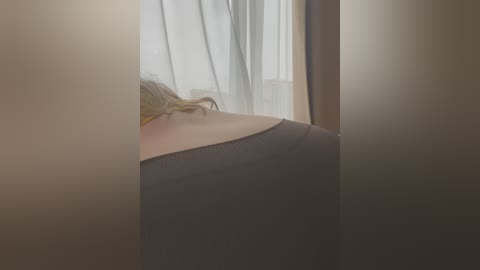 Media: Video of a blonde woman with long hair, wearing a black tank top, lying on her side, seen from behind. The background features sheer white curtains in a softly lit room.