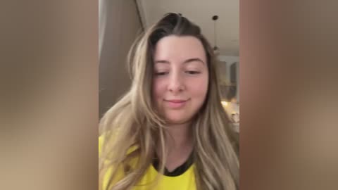 Media: Video of a young woman with long, wavy blonde hair, wearing a yellow shirt, smiling, standing in a softly lit, beige-walled room with a ceiling fan.