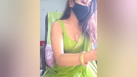 Media: Video of a South Asian woman with medium skin tone, wearing a green saree, black mask, and gold bangles, sitting indoors with a purple cushion in the background.