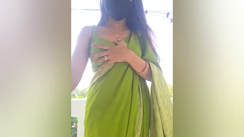 Media: Video of a woman with long dark hair, wearing a green saree and black mask, standing with her hands on her chest, against a bright, blurred outdoor background.