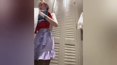 Media: Video of a young woman in a bathroom, wearing a sleeveless red top, purple skirt, and black tights, adjusting a white towel, with a beige louvered door and a white towel hanging in the background.