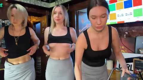 Media: Video of three young women in a bar: blonde, brunette, and a brunette with dark hair, all in black sports bras and high-waisted pants, looking down. Background includes colorful digital signage, wooden decor, and dim lighting.