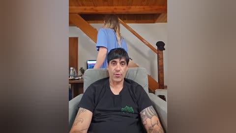 Media: Video of a man with tattoos in a black T-shirt, sitting in a chair, looking at a woman in a blue top ascending wooden stairs.