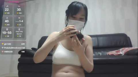 Media: Video of a young Asian woman with black hair, wearing a white sports bra, sitting on a dark brown leather couch.