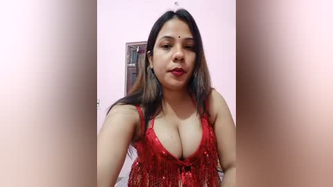 Media: Video of a South Asian woman with medium-brown skin, long black hair, and a red sequined top, showing cleavage, standing in a pink-walled room with a window and a bookshelf in the background.