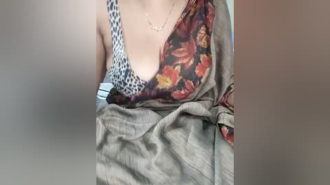 Media: A video shows a woman with fair skin, wearing a sleeveless top with a floral pattern and a gray cardigan. She has a thin gold necklace. The background is blurred.