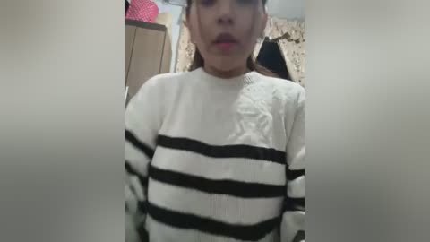 Media: A video of a young girl with light skin and straight brown hair, wearing a white and black striped sweater, making a pouty face. Background includes a pink hat, beige curtain, and wooden furniture.