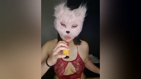 Media: Video of a woman with light skin wearing a red lace bra and a white fox mask with black ears, holding a yellow object in a dimly lit room.