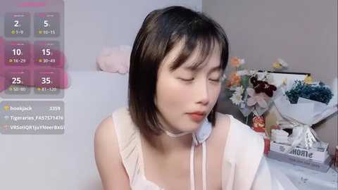 Media: Video of an East Asian woman with straight black hair, fair skin, and a petite frame, wearing a white choker and sleeveless top, sitting indoors with a floral arrangement and a weather display overlay.