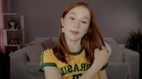 Media: A young, pale-skinned redhead girl with shoulder-length hair, wearing a yellow and green soccer jersey, poses in a cozy living room with a beige couch and bookshelves.