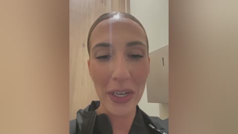 Media: Video of a woman with a sleek, high ponytail and subtle makeup, wearing a black jacket, standing in a minimalist, beige-walled bathroom with a wooden door and a towel rack.