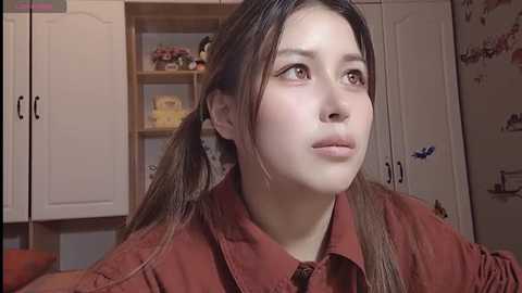 Media: Video of a young Asian woman with long brown hair in pigtails, wearing a red shirt, looking thoughtful in a cluttered bedroom with white cabinets and decorative items.