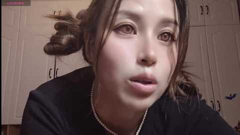 Media: Video of a young Asian woman with fair skin, brown hair in a messy bun, wearing a black top, and a pearl necklace. She has a neutral expression, and the background features white cabinets with black handles.