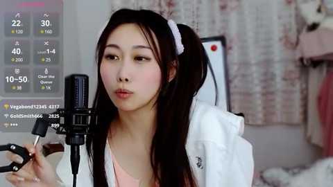 Media: Video of a young East Asian woman with long black hair in a ponytail, wearing a white lab coat, singing into a black microphone. Background shows a blurred room with a pink curtain.