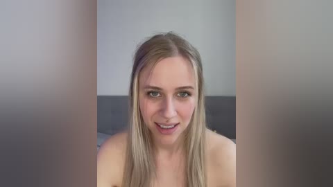 Media: Video of a smiling, topless young woman with straight blonde hair and light skin, sitting in a dimly lit room with neutral-colored walls.