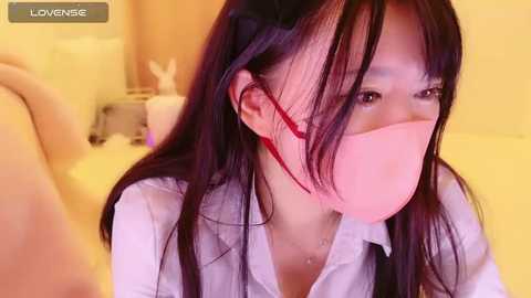 Media: A video of an Asian woman with long black hair, wearing a pink face mask and white shirt, in a softly lit bedroom.