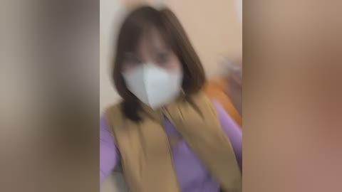 Media: Video of a person with medium-length brown hair, wearing a white mask and a beige vest over a purple shirt, standing indoors. The image is slightly blurred.