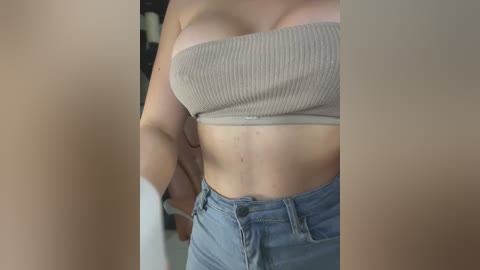 Media: Video of a light-skinned woman with medium-sized breasts, wearing a beige, ribbed tube top and blue jeans, taken from a close-up, slightly blurred perspective.