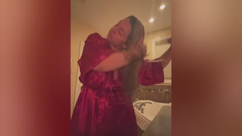 Media: Video of a young woman with long brown hair, wearing a red satin robe, standing in a bathroom with beige walls and a white sink.