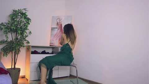 Media: Video of a woman with long, dark hair in green overalls sitting on a white chair in a minimalist, brightly lit room with a tall potted plant, white dresser, and abstract art on the wall.